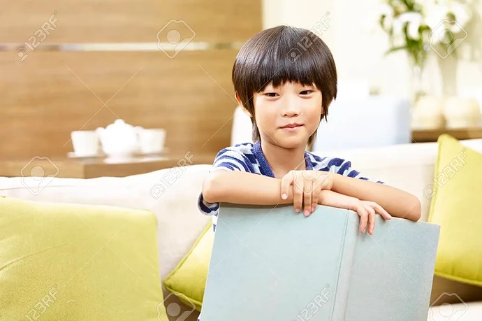 92608729-8-year-old-little-asian-boy-sitting-on-sofa-holding-a-book-looking-at-camera-smiling-