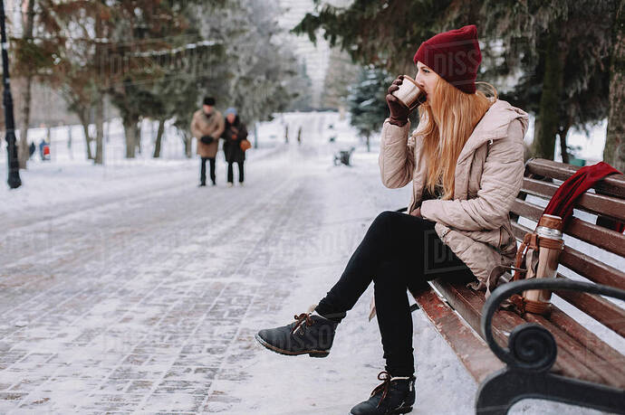 Woman drinking coffee while sitting on bench against trees at park during  winter - Stock Photo - Dissolve